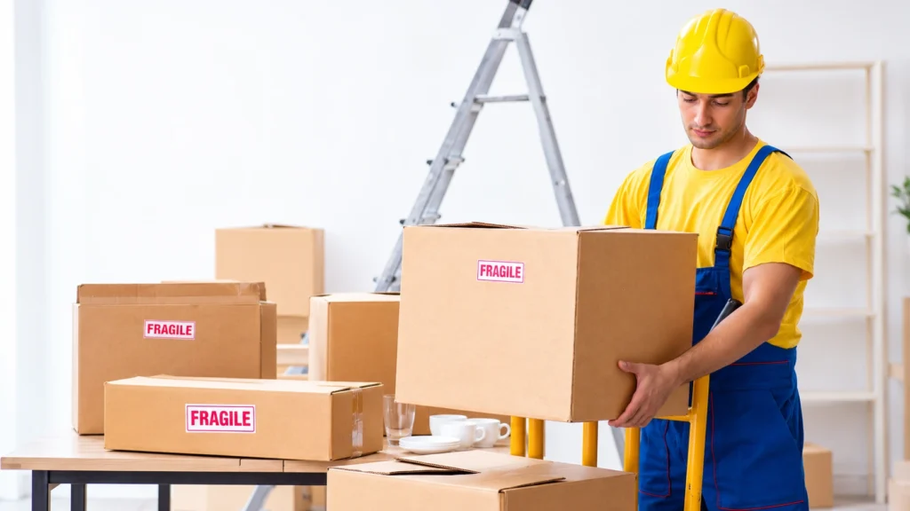 Professional mover carrying a cardboard box labeled fragile while handling packed boxes and delicate household items during a house move.
